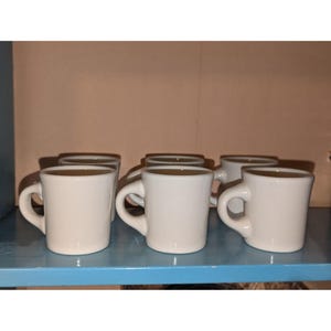 Può includere: Un set di sei tazze in ceramica bianca con manici arrotondati. Le tazze sono disposte su una mensola blu, con un muro di colore neutro sullo sfondo. Le tazze sono tutte della stessa dimensione e forma.