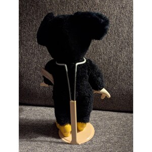 May include: A black plush toy with large ears, wearing a white-trimmed outfit and yellow shoes, is displayed on a stand. The toy has a vintage aesthetic, with a tag attached. The background is a neutral gray.