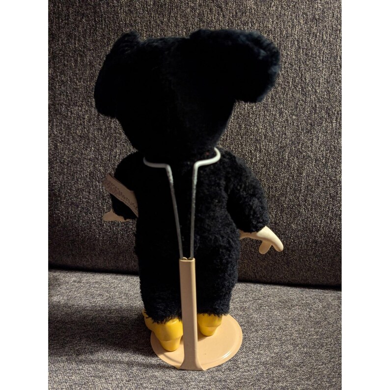 May include: A black plush toy with large ears, wearing a white-trimmed outfit and yellow shoes, is displayed on a stand. The toy has a vintage aesthetic, with a tag attached. The background is a neutral gray.