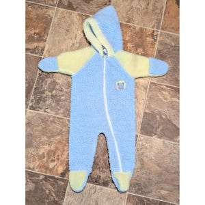 May include: A baby's blue and yellow hooded jumpsuit with a full-length zipper. The jumpsuit has a fuzzy texture and features yellow accents on the hood, sleeves, and feet. A small embroidered patch is on the chest.
