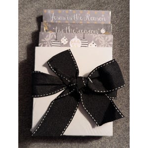 May include: A white gift box tied with a black bow with white stitching. The box is topped with three Christmas cards with text that reads "Jesus is the Reason" and "'tis the season". The cards have a gray and white design.