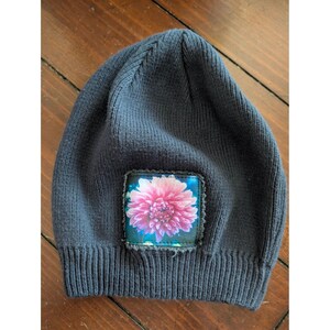 May include: A dark gray knit beanie featuring a square patch on the front. The patch displays a vibrant, detailed image of a blooming pink dahlia flower with layered petals, set against a dark blue background with a visible stitched border.