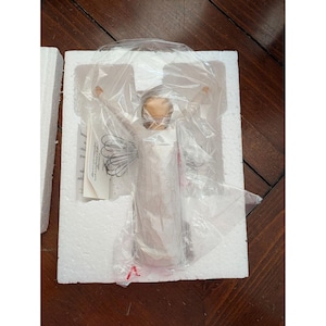 Könnte beinhalten: Eine weiße Engelsfigur mit ausgestreckten Armen, verpackt in klarem Kunststoff in einer weißen Schaumstoffform. Der Engel hat einen hellbraunen Kopf und detaillierte Flügel. Die Figur ist Teil einer Sammlerserie.