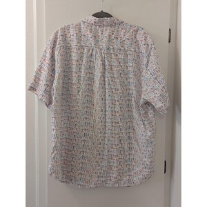 May include: A short-sleeved button-down shirt in white featuring an all-over repeating pattern of small, colorful kitchen utensils and cooking tools in shades of blue, red, and orange. The shirt has a standard collar and a straight hemline.