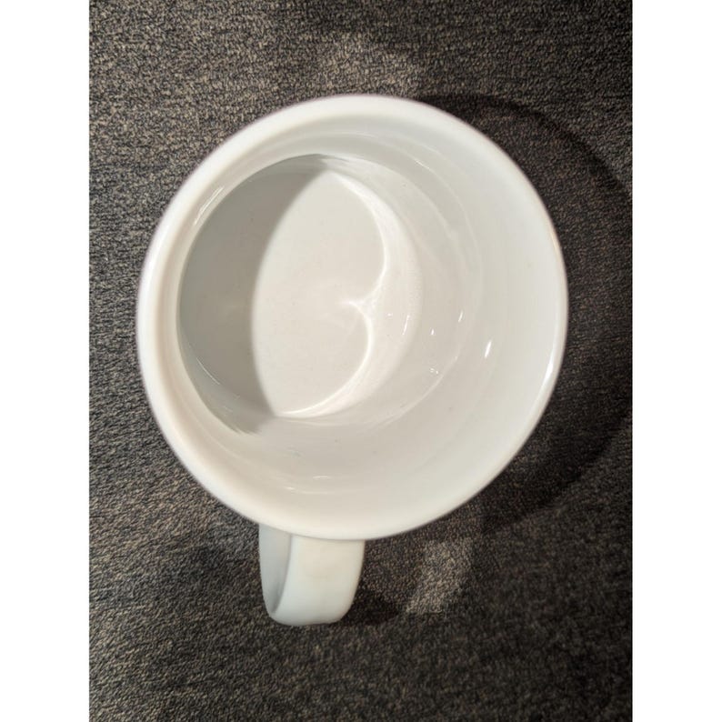 May include: A white ceramic mug with a curved handle. The mug is empty and viewed from above, showing the interior and the handle on the right side. The mug is on a dark gray surface.