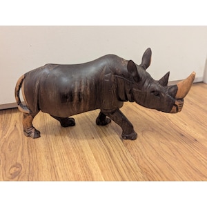 May include: A hand-carved wooden rhinoceros sculpture. The rhino is dark brown with lighter brown accents on the horn, tail, and legs. The sculpture is detailed, showing the rhino's skin texture and form. It is posed in a walking stance.