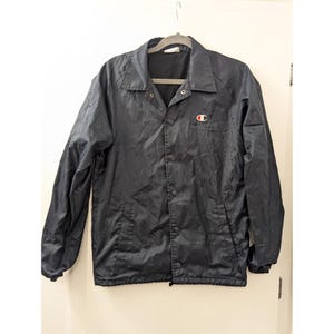 Può includere: Giacca da allenatore Champion blu navy con chiusura a bottoni e un piccolo logo rosso e bianco sul petto. La giacca è appesa a una gruccia grigia su sfondo bianco. La giacca ha un colletto e maniche lunghe.