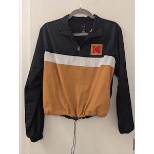 May include: Black, white, and tan windbreaker jacket with a half-zip closure and a drawstring hem. The jacket features a color-block design with a Kodak logo patch on the chest. The jacket is hanging on a gray hanger.