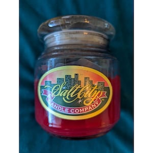 Pode incluir: Uma vela de pote de vidro com interior de cera vermelha e tampa transparente. O pote tem um rótulo com o texto "Salt City Candle Company" e um desenho de paisagem urbana em amarelo, laranja e preto.