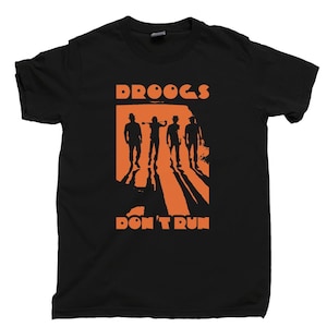 May include: Black t-shirt featuring an orange graphic with the text "DROOGS DON'T RUN." The graphic depicts four silhouetted figures with shadows, standing in front of an orange rectangle. The shirt is made of a soft material.