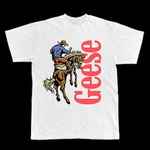 Op de afbeelding: Wit t-shirt met een afbeelding van een cowboy op een wild paard. De cowboy draagt een blauw overhemd en een bruine hoed. Het woord "Geese" is verticaal in het rood afgedrukt aan de rechterkant van het shirt.
