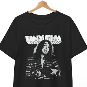 Op de afbeelding: Zwart T-shirt met een zwart-wit afbeelding van Tiny Tim, en de naam "TINY TIM" in witte blokletters boven de afbeelding. De afbeelding toont Tiny Tim in een geruite jas.