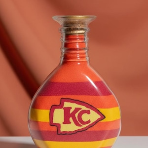 Op de afbeelding: Een decoratieve glazen fles gevuld met lagen oranje, rood en geel zand. De fles heeft het logo van de Kansas City Chiefs. De fles heeft een kurk en wordt gepresenteerd tegen een oranje achtergrond.