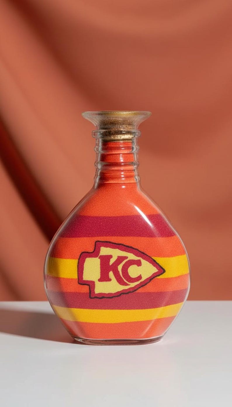 Op de afbeelding: Een decoratieve glazen fles gevuld met lagen oranje, rood en geel zand. De fles heeft het logo van de Kansas City Chiefs. De fles heeft een kurk en wordt gepresenteerd tegen een oranje achtergrond.