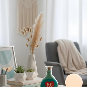 May include: A decorative vignette featuring a teal and red heart-shaped bottle with "KC" text, a white vase with dried flowers, and a small succulent plant. A glowing orb lamp and a macrame wall hanging complete the scene.