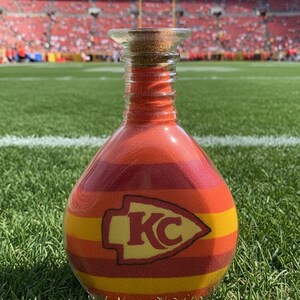 Op de afbeelding: Een decoratieve glazen fles gevuld met lagen rood, oranje en geel zand. De fles heeft het logo van de Kansas City Chiefs en een kurk. De achtergrond toont een voetbalstadion.