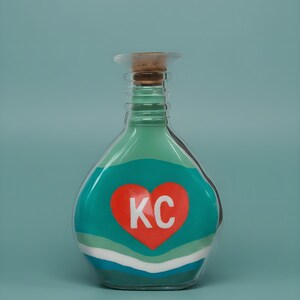 May include: A clear glass bottle with a cork stopper, filled with layers of colored sand. The bottle has a teal and white design with a red heart containing the letters "KC". The background is a solid teal color.