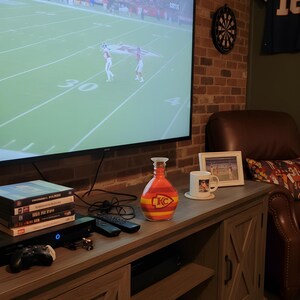 Op de afbeelding: Een entertainmentcenter in de woonkamer met een grote televisie waarop een American football-wedstrijd te zien is. Een decoratieve oranje en gele fles met een "KC"-logo staat op de console, samen met dvd's, afstandsbedieningen en een gameconsole.