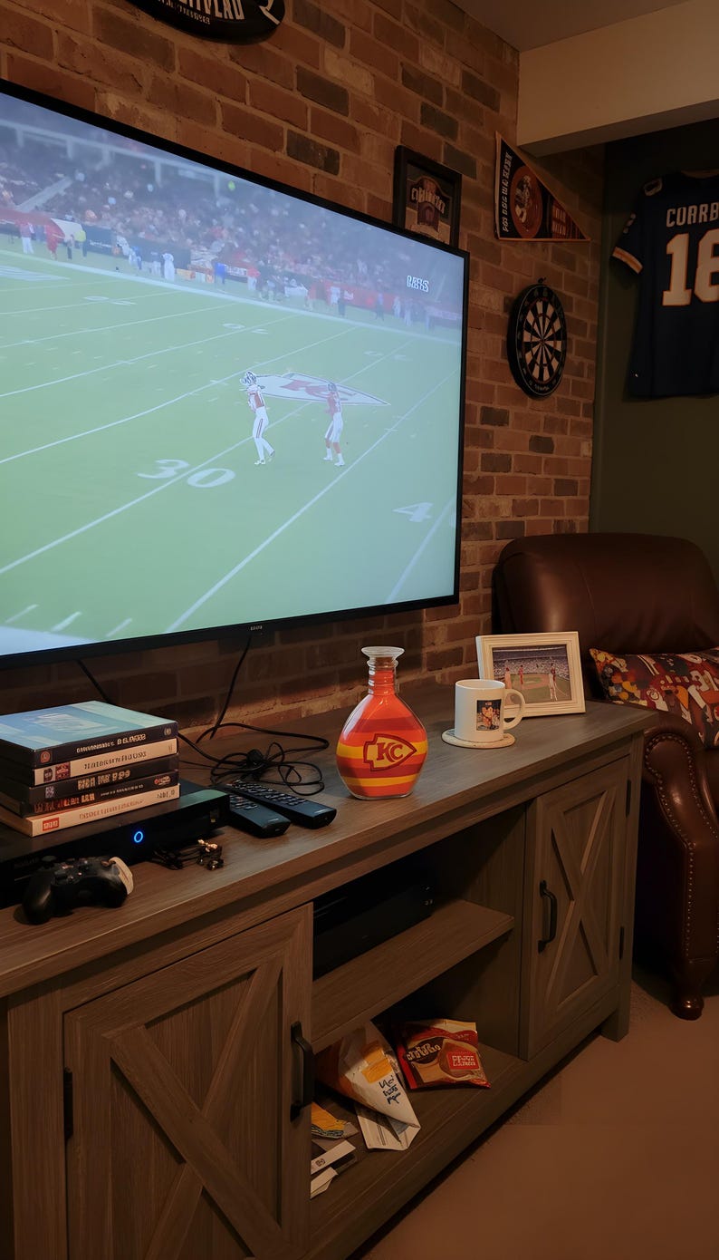 Op de afbeelding: Een entertainmentcenter in de woonkamer met een grote televisie waarop een American football-wedstrijd te zien is. Een decoratieve oranje en gele fles met een "KC"-logo staat op de console, samen met dvd's, afstandsbedieningen en een gameconsole.