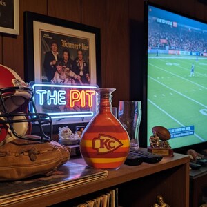 Op de afbeelding: Een decoratieve vaas met een rood, geel en oranje ontwerp, met het logo van de Kansas City Chiefs. De vaas staat op een plank met een voetbal, een ingelijste foto en een neonbord met de tekst "THE PIT".