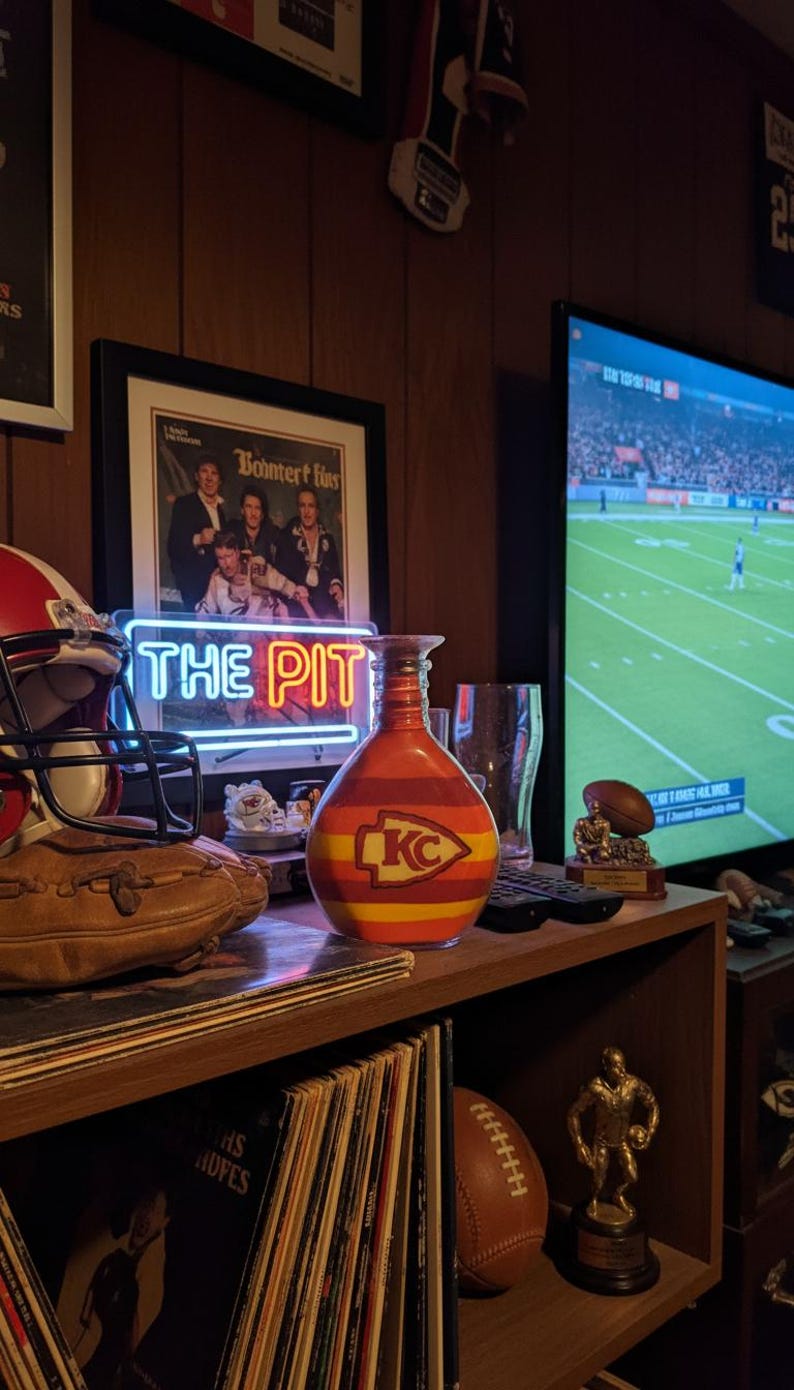Op de afbeelding: Een decoratieve vaas met een rood, geel en oranje ontwerp, met het logo van de Kansas City Chiefs. De vaas staat op een plank met een voetbal, een ingelijste foto en een neonbord met de tekst "THE PIT".