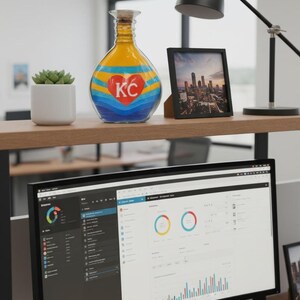 K&ouml;nnte beinhalten: Eine dekorative Glasflasche mit Korken, mit gelbem Oberteil, blauem und rotem Herzdesign und den Buchstaben "KC". Ein gerahmtes Stadtlandschaftsfoto, eine Schreibtischlampe und ein Computermonitor sind ebenfalls sichtbar.