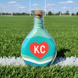 May include: A decorative glass bottle filled with layers of sand in shades of blue, white, and green. A red heart with the letters "KC" is centered on the bottle. The bottle has a cork stopper and is set on a green field.