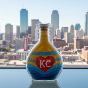 K&ouml;nnte beinhalten: Eine dekorative Glasflasche gef&uuml;llt mit Schichten aus gelbem, blauem und t&uuml;rkisfarbenem Sand. Ein rotes Herz mit "KC" in Wei&szlig; ist auf der Flasche zentriert. Der Hintergrund zeigt eine Stadtlandschaft mit hohen Geb&auml;uden.