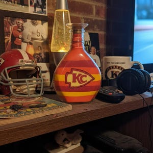 Op de afbeelding: Een decoratieve glazen fles met een rood, geel en oranje gestreept ontwerp, met het logo van de Kansas City Chiefs. De fles staat op een houten plank met een voetbalhelm en andere memorabilia.