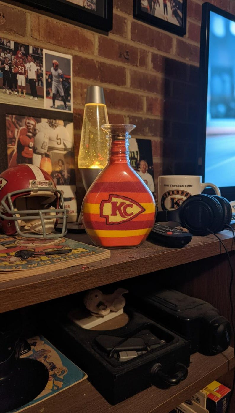 Op de afbeelding: Een decoratieve glazen fles met een rood, geel en oranje gestreept ontwerp, met het logo van de Kansas City Chiefs. De fles staat op een houten plank met een voetbalhelm en andere memorabilia.