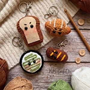 Puede incluir: Cuatro llaveros de comida hechos a mano con ganchillo: una rebanada de pan tostado, un cruasán, un rollo de sushi y un éclair de chocolate. Cada llavero tiene un cierre de metal. Los llaveros están sobre una superficie de madera con hilo y suministros de ganchillo.
