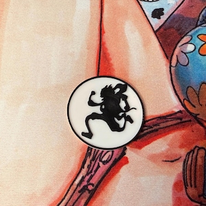 May include: A round, white button with a black silhouette of a character in motion. The button has a black border and is set against a colorful background with cartoon figures and floral patterns.