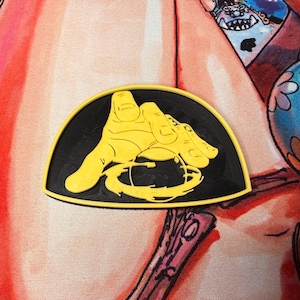 May include: A black and yellow emblem featuring a hand holding a circular object. The hand is yellow, and the background is black. The emblem is in a semi-circular shape.