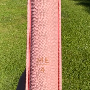 Op de afbeelding: Een roze boek met de tekst "ME 4" in reli&euml;f op de kaft. Het boek heeft een gestructureerd oppervlak en een afgeronde rug. De achtergrond is groen gras.
