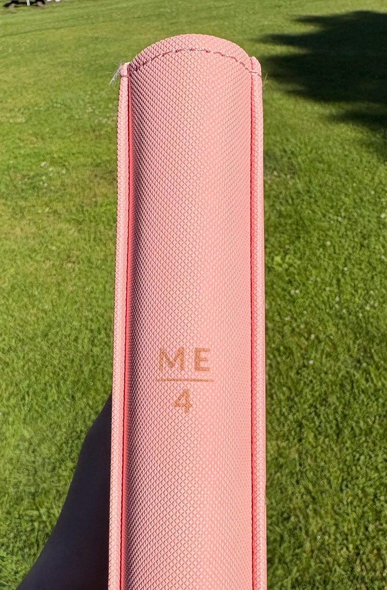 Op de afbeelding: Een roze boek met de tekst "ME 4" in reli&euml;f op de kaft. Het boek heeft een gestructureerd oppervlak en een afgeronde rug. De achtergrond is groen gras.