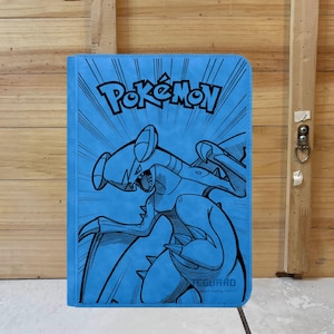 Puede incluir: Un archivador azul para cartas Pokémon con una ilustración negra de un personaje Pokémon. El logotipo de Pokémon está en la parte superior, con un fondo de estilo cómic. El archivador es rectangular y parece estar hecho de un material texturizado.