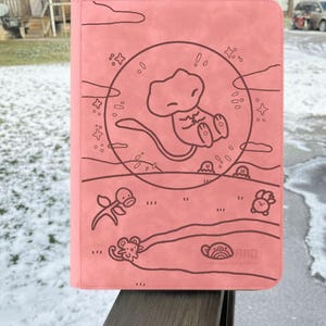 Puede incluir: Un archivador de tarjetas rosa con un dibujo lineal marrón de un personaje de dibujos animados dentro de un círculo, con otros personajes y formas de dibujos animados. El archivador se sostiene al aire libre en un día de nieve.