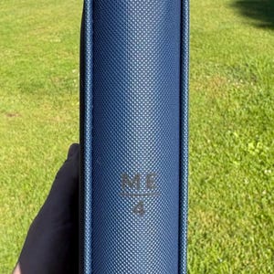 Op de afbeelding: Een marineblauw, getextureerd boek of map met de letters "ME" en het nummer "4" in reli&euml;f op de omslag. De rug is zichtbaar en het item wordt tegen een achtergrond van groen gras gehouden.