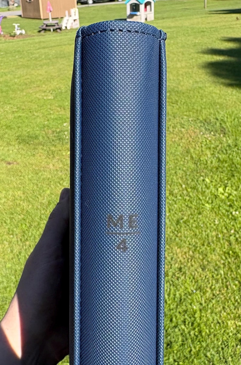 Op de afbeelding: Een marineblauw, getextureerd boek of map met de letters "ME" en het nummer "4" in reli&euml;f op de omslag. De rug is zichtbaar en het item wordt tegen een achtergrond van groen gras gehouden.