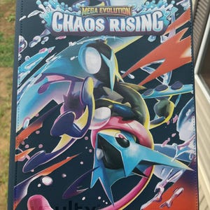 Peut inclure: Un classeur de cartes à collectionner rectangulaire avec un fond sombre et un graphique coloré représentant une créature bleue et noire. Le texte MEGA EVOLUTION CHAOS RISING est imprimé en blanc et bleu en haut. Le design inclut des éclaboussures d'eau et des traînées d'énergie orange.