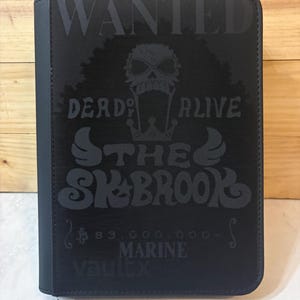 May include: Black zippered binder featuring a skull graphic with afro hair and the words "WANTED DEAD OR ALIVE THE SKBROOK" in grey. The binder also has the text "83,000,000 MARINE vaultx".