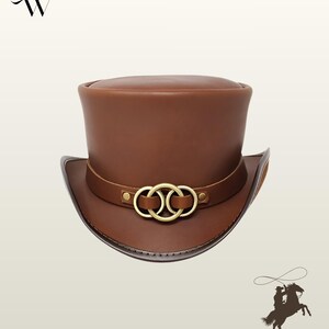 May include: A brown leather top hat featuring a flat crown and a curved brim. A leather band encircles the base of the crown, adorned with three interlocking brass-toned metal rings secured by small rivets on each side of the decorative hardware.