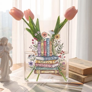 May include: A clear rectangular glass vase with a stack of illustrated books and flowers. The vase holds pink tulips and other greenery. The books are decorated with floral designs. A white angel statue is in the background.