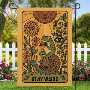 May include: A garden flag with a whimsical design featuring a green frog playing a flute while riding a snail. The flag has a yellow background with floral and eye motifs, and the words "STAY WEIRD".