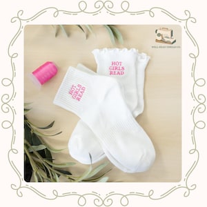 May include: A pair of white crew socks featuring a ruffled top edge and the embroidered pink text "HOT GIRLS READ" on the ankle. The socks are positioned next to a spool of bright pink thread and a small logo for Well-Read Thread Co.