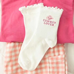 May include: A pair of white ribbed socks with ruffled cuffs, featuring an embroidered pink cowboy hat icon and the text "COWBOY LOVER" in pink lettering on the side of each sock, positioned against a bright pink fabric and a peach gingham fabric.