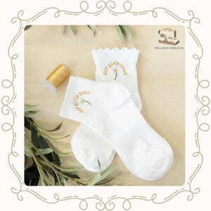 May include: A pair of white ribbed ankle socks with ruffled cuffs. Each sock features an embroidered daisy flower with the text "hot girls read" arched above it in gold thread. A spool of gold thread is positioned next to the socks.