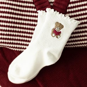May include: A pair of white ribbed socks featuring a ruffled top edge and an embroidered brown teddy bear holding a small red book. The socks are crafted with a soft texture and display a detailed stitched design of the bear on the upper ankle.