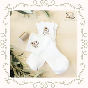 May include: A pair of white ribbed ankle socks with ruffled cuffs, each featuring a small brown embroidered cowboy figure holding a lasso. The socks are displayed alongside a spool of thread and a logo reading WELL-READ THREAD CO.