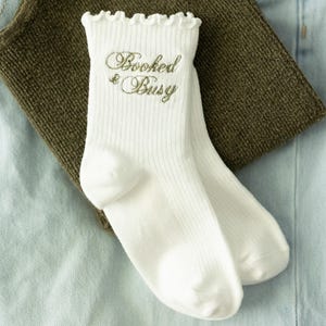 May include: A pair of white ribbed crew socks featuring a ruffled top edge and the text "Booked & Busy" embroidered in a metallic gold script font on the upper portion of the sock. The socks have a classic knit texture and a clean, simple design.